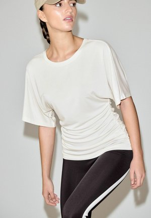 Jasnobeżowa bluzka z krótkim rękawem o luźnym kroju, z marszczeniem po bokach, zestawiona z czarnymi legginsami z wysokim stanem, ozdobionymi białymi paskami.