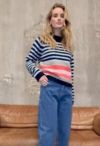 Pull rayé bleu marine, crème, rose et rouge, avec une finition texturée. Associé à un jean bleu clair à coupe décontractée. Porté devant un mur texturé.