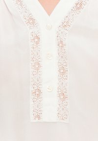 Blouse blanche devant avec un col en V, trois boutons et une décoration en dentelle le long de la patte de boutonnage.