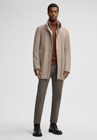Man met een beige wollen jas over een bruine coltrui, grijze broek en zwarte veterschoenen, staande tegen een witte achtergrond.