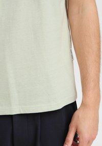 Cămașă scurtă, de culoare verde deschis, cu o textură netedă, având un etichetă la tiv inscripționată cu "VINERI", purtată împreună cu pantaloni negri.