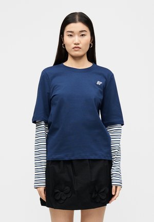 Žena s dlhými rovnými čiernymi vlasmi, ktorá nosí zlaté kruhové náušnice, námornícky modrú krátkym rukávom košeľu cez pruhovaný dlhý rukáv top a čiernu sukňu s kvetinovými detailmi.