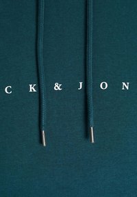 Teal hettegenser med hvit tekst "CK&JON." Har flettede snorer med sølvstopper. Glatt, ubehandlet teksturoverflate.