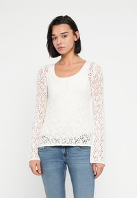 Λευκή δαντελένια μπλούζα με μακριά μανίκια και στρογγυλή λαιμόκοψη. Διαθέτει υφή με μοτίβο και εφαρμοστή γραμμή, συνδυασμένη με μπλε τζιν.