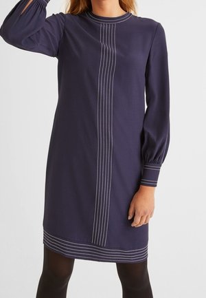 Tmavě modré šaty s dlouhým rukávem, bílé svislé a vodorovné švy, rukávy s manžetami, nošené s černými punčochami.