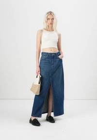 Denim maxi skirt with a front slit, light blue color, high waist, paired with a fitted white crop top and a small woven bag.