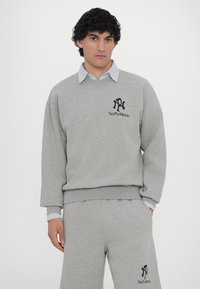 Pulover gri și pantaloni de trening asortați dintr-un material moale, având un logo negru cu textul "No Problemo." Stratificat cu o cămașă albastră cu guler.