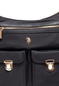 Sort nylon taske med guldfarvet hardware, to frontlommer med tryklåse, en lynlås i toppen og et logoaccent. Glat tekstur og struktureret design.