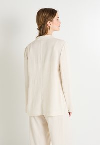 Blazer de lino color beige claro con una forma estructurada, mangas largas y una textura suave. Presenta una costura trasera sutil y un diseño minimalista.