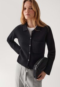 Giovane donna che indossa un cardigan nero a coste con bottoni sopra una maglietta bianca, mentre tiene una piccola borsa nera e dei pantaloni grigi con coulisse su uno sfondo neutro.