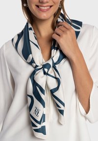 Foulard en soie avec un motif abstrait bleu marine et crème, noué autour du cou, avec un petit logo dans un coin. Texture douce et lisse.