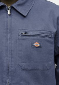 Κοντινό πλάνο ενός μπλε μπουφάν Dickies με τσέπη στο στήθος με φερμουάρ και μεταλλικό τράβηγμα φερμουάρ στο μπροστινό κλείσιμο.