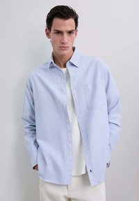 Camicia azzurra chiaro a righe sottili, con maniche lunghe, una tasca sul petto e colletto, indossata sopra una maglietta bianca.