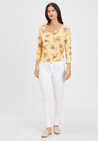 Gelbes Blumenmuster Langarmshirt mit V-Ausschnitt und Knopfverschluss, kombiniert mit weißen Slim-Fit-Jeans und beigen durchlöcherten Slippern.