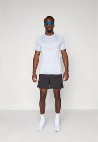 Šviesiai mėlynas sportinis marškinėlis su tekstūruotu raštu, derinamas su juodomis šortais. Modelis dėvi mėlynas saulės akinius ir baltus sportinius batelius.