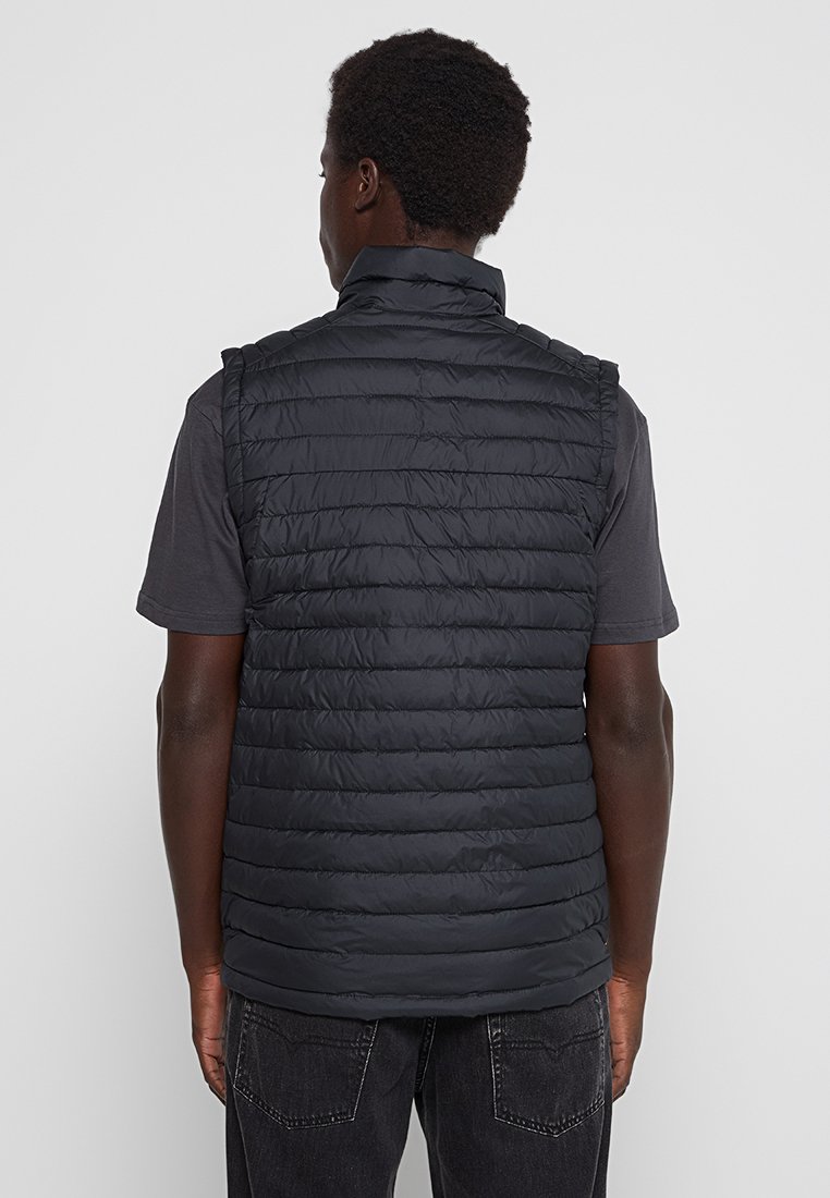 Uomo che indossa un gilet quilted senza maniche nero e una maglietta scura, in piedi rivolto di spalle contro uno sfondo chiaro uniforme.