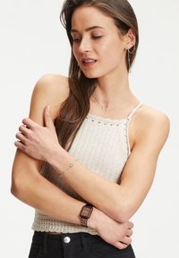 Braune quadratische Uhr mit Metallarmband, silberne Kettenarmband mit kreisförmigen Akzenten und einem beigen Stricktop mit gewelltem Saum.