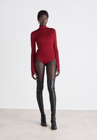 Červený bodysuit s dlhými rukávmi a vysokým golierom, v kombinácii s priesvitnými čiernymi pančuchami s bodkami a čiernymi čižmami po stehná, čím vytvára vrstvený vzhľad.