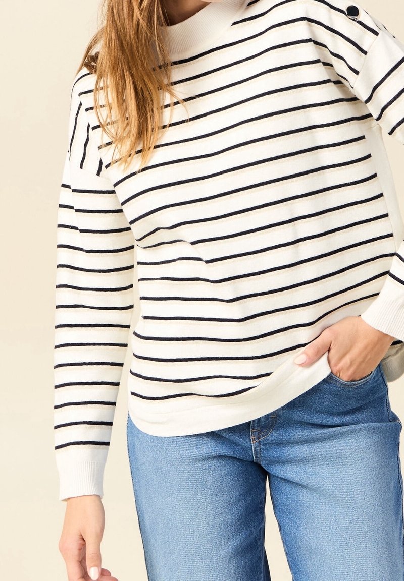 Striped sweater in cream with navy stripes, featuring a round neckline and button accents on the shoulder. Worn with blue jeans.