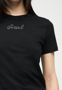 Μαύρο κοντομάνικο t-shirt με μικρό λογότυπο "diesel" σε στυλ κρυστάλλων κοντά στον γιακά, φορεμένο από άτομο με ορατό λαιμό και χέρι.