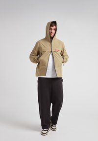 Chaqueta caqui con cremallera en el frente, capucha y bolsillos laterales; combinada con pantalones negros holgados y zapatillas bicolores. Presenta un logo de marca rojo.