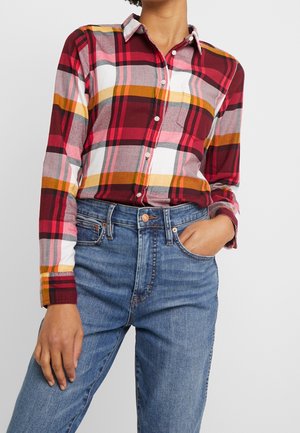 Kvinde iført rød, orange, hvid og lyserød ternet skjorte med knapper, stoppet ned i højtaljede blå jeans, med den ene hånd i lommen.