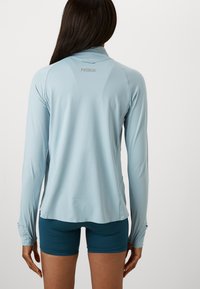 Light blue long-sleeve athletic shirt with a high collar, side slits at the hem, and logo detail on the upper back. Smooth texture.