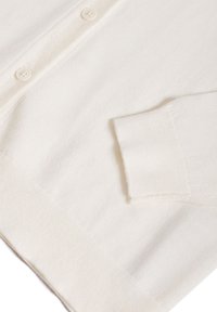 Bílý svetr s knoflíkovou přední částí, žebrovanými manžetami a měkkou strukturou. Obsahuje jednu kapsu a má volnější střih.