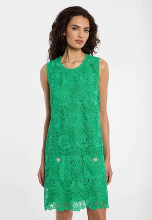 Vrouw met donker krullend haar die een mouwloze groene kanten jurk draagt met bloemmotieven en decoratieve parelknopen op de voortassen.