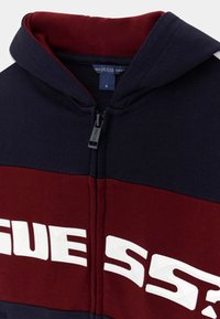 Bordeauxrode en marineblauwe hoodie met ritssluiting, voorzien van een wit logo op de borst, met een zachte textuur en een capuchon. Ritsdetail zichtbaar bij de halslijn.