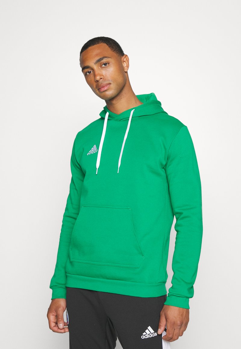 Sweat à capuche vert avec une poche frontale, un cordon de serrage et des accents blancs. Présente le logo Adidas sur la poitrine. Le matériau semble lisse et extensible.