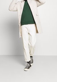 Persona che indossa un cappotto beige, una felpa verde, pantaloni bianchi e sneakers alte nere mentre cammina su uno sfondo neutro.