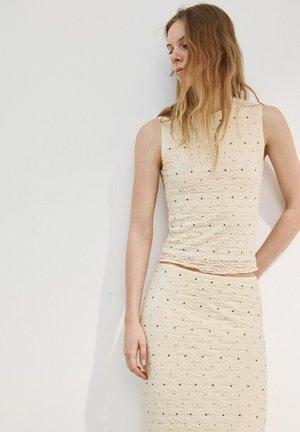 Jeune femme aux longs cheveux blonds portant un haut sans manches crème texturé et une jupe assortie avec petits points noirs sur un fond uni.