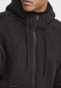 Black fleece hoodie with a textured, fluffy surface. Features a full zip closure, adjustable drawstrings, and a hoodie neckline.
