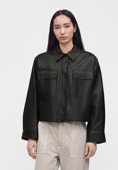 Young woman with long dark hair wearing a black buttoned leather jacket and light beige pants, standing against a plain white background.