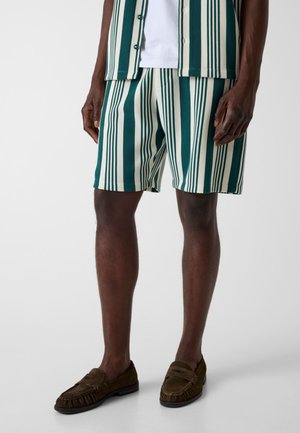 Man draagt groen-witte vertical gestreepte korte broek, bijpassend T-shirt met korte mouwen, wit ondershirt en bruine slip-on loafers.