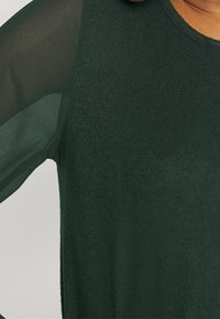 Dunkelgrünes Oberteil mit strukturiertem Strickkörper und transparenten, leichten Ärmeln. Der Halsausschnitt ist rund, und der Stoff wirkt weich und glatt.