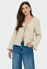 Beige, strukturierte Bluse mit langen Ärmeln, V-Ausschnitt und vorderem Bindedetail. Kombiniert mit hochgeschnittenen blauen Jeans.