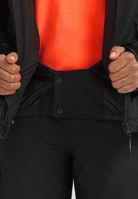 Zwarte jas met een contrasterende oranje binnenvoering, met een drukknoopsluiting op de taille en een gestructureerde stof. Handen grijpen naar de randen van de jas.