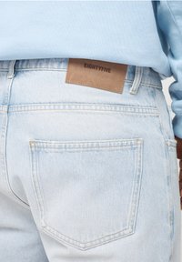 Jasnoniebieskie dżinsy o gładkiej fakturze, z brązową skórzaną metką z napisem "EIGHTYFIVE" na pasku oraz standardowym wzorem tylnej kieszeni.