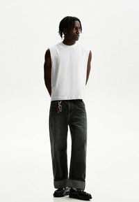 Hvid ærmeløs T-shirt parret med løse, mørkegrå denimjeans. Bemærkelsesværdig detalje: nøglekæde-accessory i baglommen. Fodtøj: sorte sko.