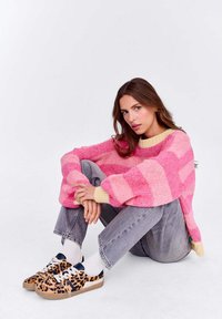 Przesadnie duży różowy sweter z dzianiny z żółtymi akcentami, połączony z szarymi dżinsami i trampkami w print w panterę. Modelka siedzi, opierając ramiona na kolanach.