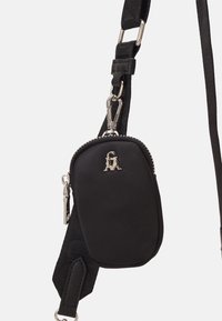 Svart kompakt crossbody-påse med silverblixtlås och logotyp, fäst vid justerbar svart tygrem med metallspännen.