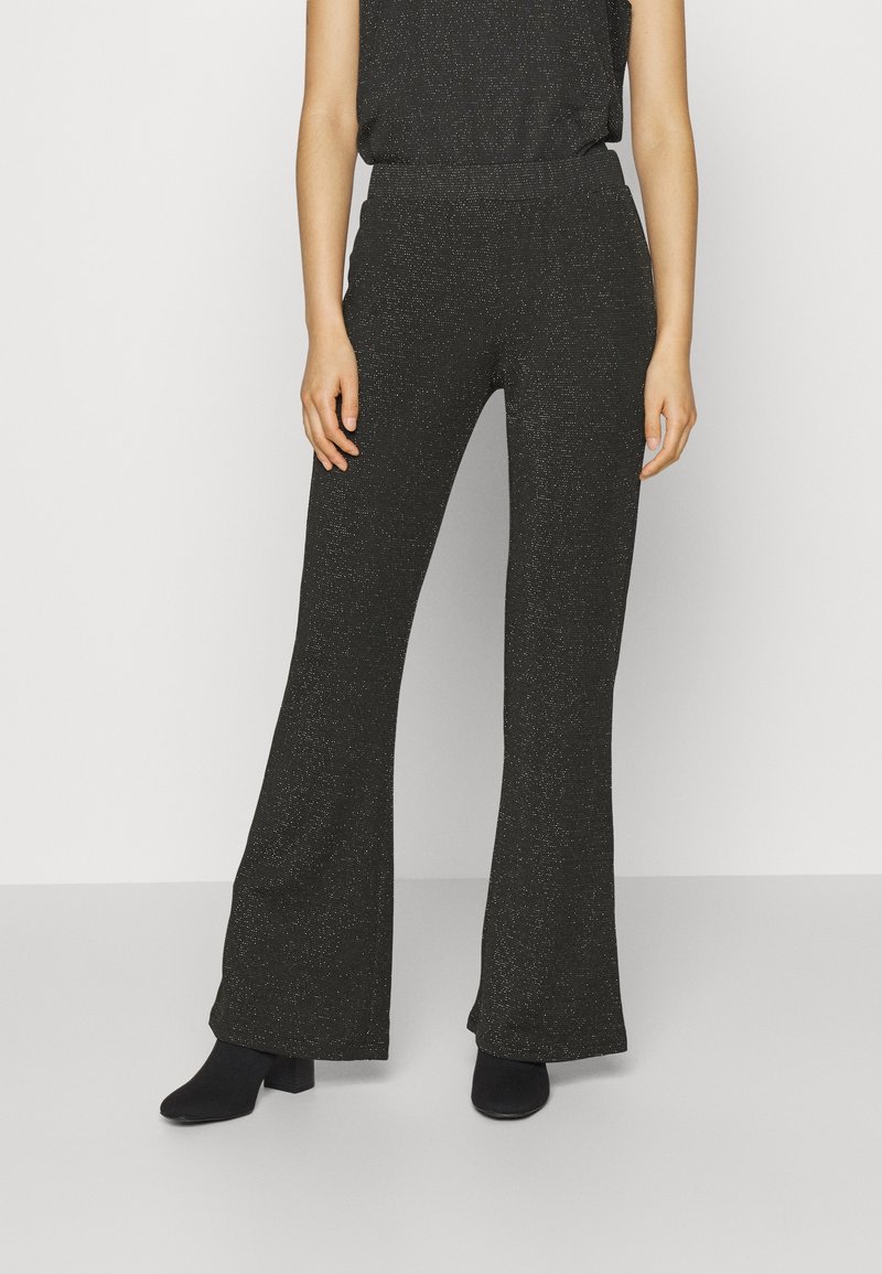 Femme portant un pantalon large noir pailleté et des bottines noires à talons, debout devant un fond clair uni.
