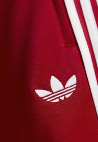 Κόκκινο ύφασμα με λευκό λογότυπο Adidas trefoil και τρεις λευκές κάθετες ρίγες δίπλα σε ραφή τσέπης.
