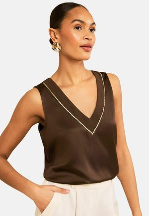 Mujer con blusa marrón sin mangas y escote en V con costuras claras, grandes pendientes dorados y pantalones blancos, con la mano en el bolsillo, mirando hacia la derecha.