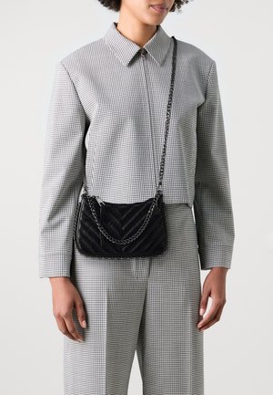 Kobieta ubrana w czarno-białą kratowaną kurtkę i spodnie, niosąca czarną pikowaną torebkę na ramię z łańcuszkowym paskiem.
