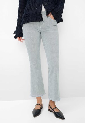 Frau trägt hellblaue und weiß gestreifte, gekürzte Hose mit ausgefransten Säumen, einen schwarzen, strukturierten Pullover mit Fransen und schwarze, spitze Slingback-Schuhe.