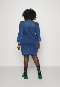 Femme aux cheveux bouclés portant une robe en jean et des bottines noires avec des semelles vertes, debout face à un mur uni gris clair.