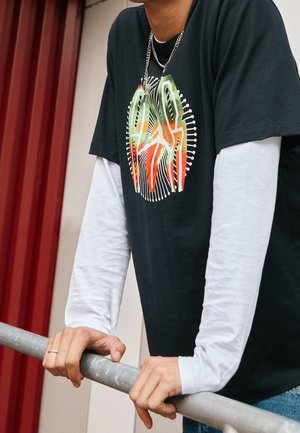 Schwarzes Grafik-T-Shirt, das über einem weißen Langarmshirt getragen wird. Das T-Shirt hat ein farbenfrohes Design mit Linien und kreisförmigen Akzenten.